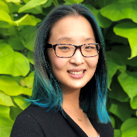 headshot of Catherine Kim, Asian girl with glasses, blue hair in front of a green leafy wall